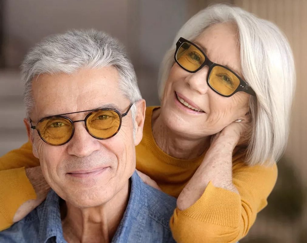 Ein fröhliches älteres Paar posiert zusammen, lächelnd und einen warmen Moment teilend, gekleidet in Freizeitkleidung vor einem sanft verschwommenen Hintergrund.