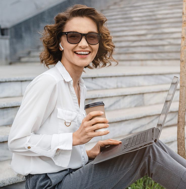 Eine Frau mit Sonnenbrille sitzt auf einer Bank und arbeitet an einem Laptop in einer sonnigen Umgebung im Freien